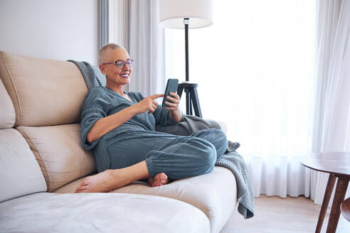 Smiling woman using smart phone while relaxing on sofa at home - AGOF00162