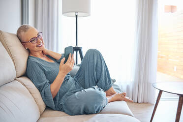 Lächelnde reife Frau, die ein Smartphone benutzt, während sie zu Hause auf dem Sofa sitzt - AGOF00164