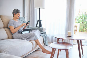 Frau, die ihr Smartphone benutzt, während sie auf dem Sofa im heimischen Wohnzimmer sitzt - AGOF00171
