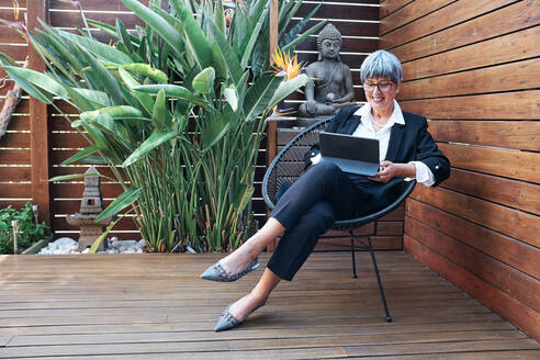 Reife Geschäftsfrau, die ein digitales Tablet benutzt, während sie auf einem Stuhl im Hinterhof sitzt - AGOF00178