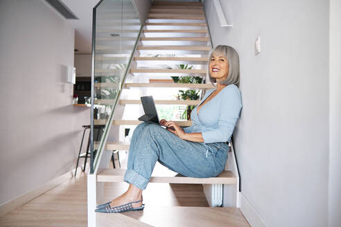 Mature woman with digital tablet sitting on steps at home - AGOF00185