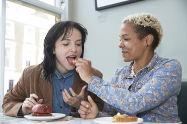 Smiling woman feeding food to girlfriend sitting in cafe - PMF01917