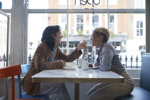 Happy young woman feeding food to girlfriend sitting in cafe - PMF01923