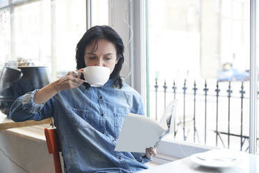 Junge Frau trinkt Kaffee, während sie in einem Café sitzt und ein Buch liest - PMF01928