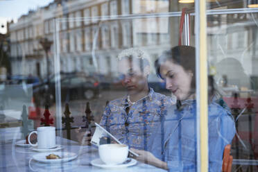 Junge Frau, die mit ihrer Freundin im Café sitzt und ein Buch liest, gesehen durch ein Glasfenster - PMF01931