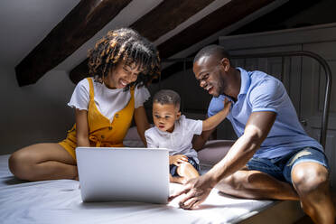 Happy family with son sitting on bed using laptop in attic at home - OCMF02151