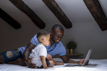 Father using laptop with son on bed in attic at home - OCMF02152