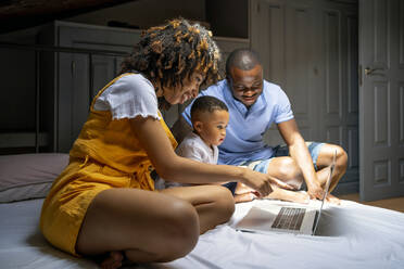 Multi-ethnic family with laptop sitting on bed in attic at home - OCMF02157