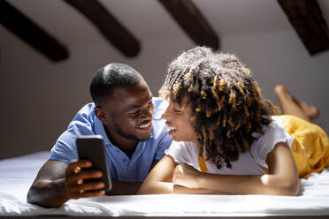 Lächelnder Mann mit Smartphone, der eine Frau ansieht, während er zu Hause auf dem Bett liegt - OCMF02158