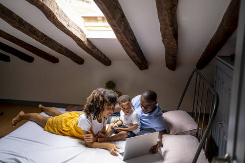 Father and mother with son using laptop on bed in attic at home - OCMF02159