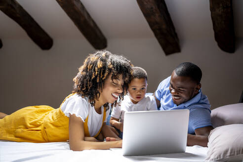 Lächelnde Familie, die einen Laptop mit ihrem Sohn benutzt, während sie zu Hause auf dem Bett liegt - OCMF02161