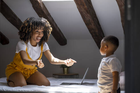 Excited mother with laptop calling son while sitting on bed at home - OCMF02164