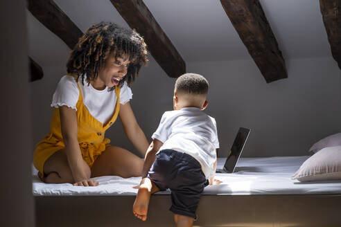 Mother looking at son climbing on bed at home - OCMF02166