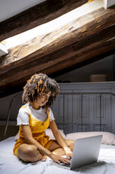 Afro woman using laptop while sitting on bed in attic at home - OCMF02168