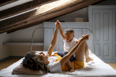 Happy mother playing with son while lying on bed in attic at home - OCMF02179