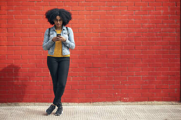 Young woman using smart phone while standing in front of red brick wall - JSRF01554