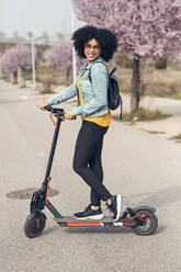 Smiling young woman standing with electric push scooter on road - JSRF01556