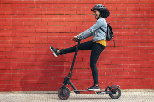 Carefree woman riding electric push scooter by red brick wall - JSRF01562