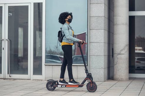 Woman with protective face mask riding electric push scooter by building during COVID-19 crisis - JSRF01563