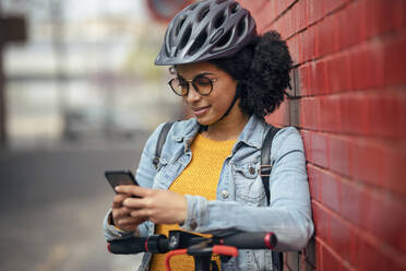 Young woman with electric push scooter using smart phone while leaning on brick wall - JSRF01564