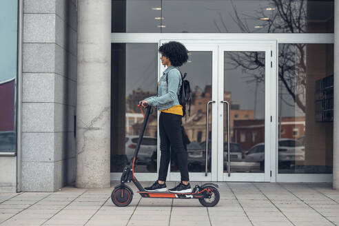 Woman riding electric push scooter on footpath near building - JSRF01565