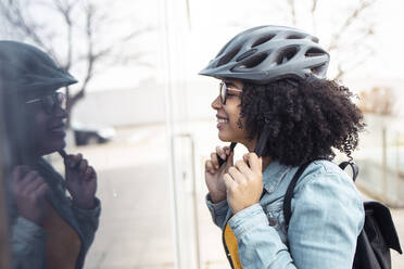 Lächelnde junge Frau, die ein Glasfenster betrachtet und einen Fahrradhelm trägt - JSRF01566