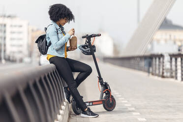 Afro-Frau mit Elektroroller isst Essen, während sie sich auf ein Geländer stützt - JSRF01569