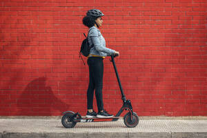 Young woman with backpack riding electric push scooter by red wall - JSRF01576