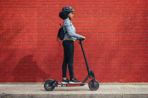 Young woman with backpack riding electric push scooter by red wall - JSRF01576