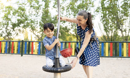 Smiling grandmother supporting grandson playing at playground in park - JCCMF02966