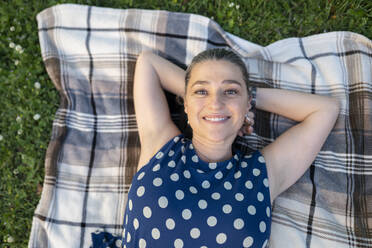 Lächelnde reife Frau mit Händen hinter dem Kopf auf einer Decke im Park liegend - JCCMF02977