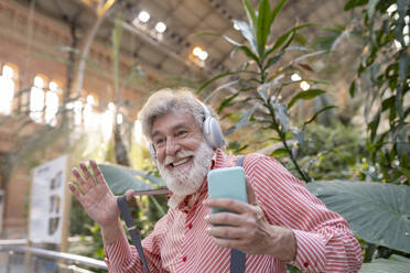 Happy white bearded mature man dancing while listening music in front of plants - JCCMF02981