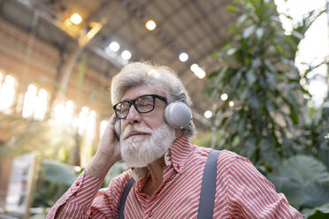 Älterer Mann mit weißem Haar und Brille hört Musik über drahtlose Kopfhörer am Bahnhof - JCCMF02983