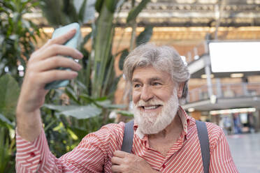 Happy white bearded man taking selfie through smart phone at station - JCCMF02988