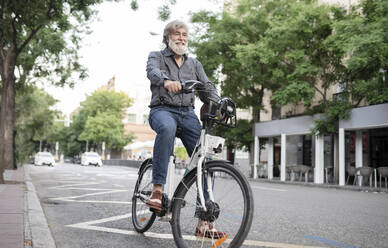 Happy mature man with electric bicycle on street in city - JCCMF03006