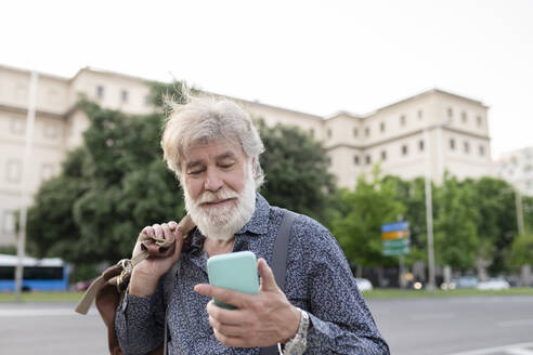 White bearded man carrying luggage while looking at smart phone in city - JCCMF03008