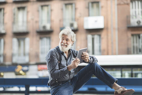 Smiling mature man looking away while sitting with digital tablet in city - JCCMF03016