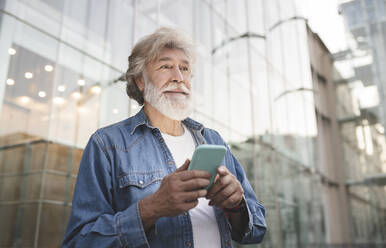 Älterer Mann, der sein Smartphone hält und vor einem Glasgebäude wegschaut - JCCMF03022