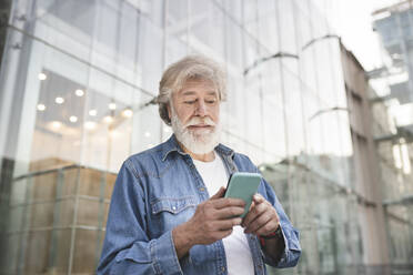 Weißer bärtiger Mann mit Smartphone vor einem modernen Glasgebäude - JCCMF03023