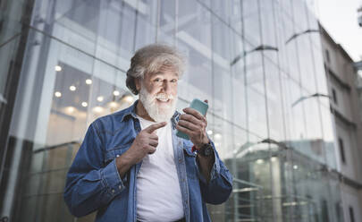 Glücklicher reifer Mann, der auf sein Smartphone vor einem Glasgebäude zeigt - JCCMF03024