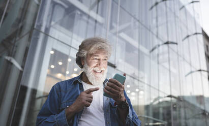Glücklicher reifer Mann, der auf sein Smartphone vor einem modernen Glasgebäude zeigt - JCCMF03026