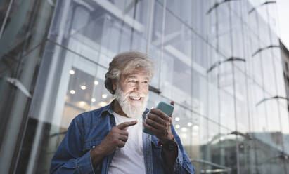 Happy mature man pointing at smart phone in front of modern glass building - JCCMF03026