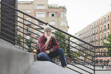 Mature man wearing knit hat sitting with hand on chin at staircase - JCCMF03029