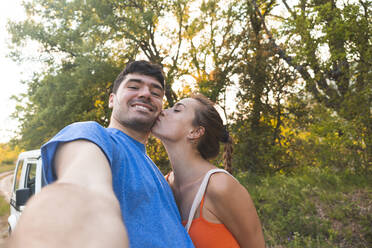 Woman kissing boyfriend while taking selfie at forest - JAQF00682