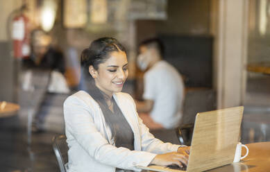 Weibliche Fachkraft, die einen Laptop benutzt, während sie in einem Café sitzt, gesehen durch ein Fensterglas - JCCMF03042