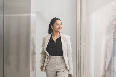 Smiling businesswoman holding laptop while walking at office - JCCMF03044