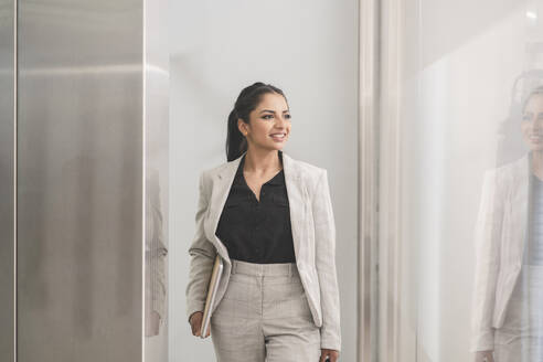 Lächelnde Geschäftsfrau hält Laptop beim Gehen im Büro - JCCMF03044