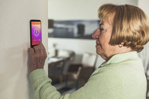 Senior woman using smart thermostat on wall at home - UUF23654