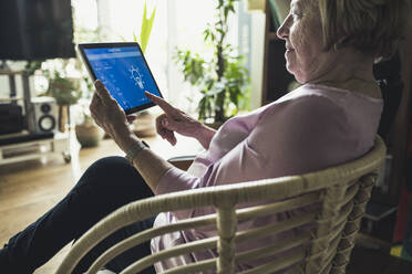 Senior woman checking smart home device while sitting on chair - UUF23670