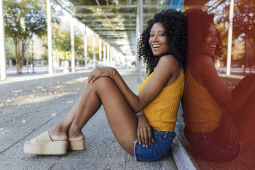Junge Frau lacht, während sie auf dem Fußweg vor einer roten Glaswand sitzt - JRVF01162